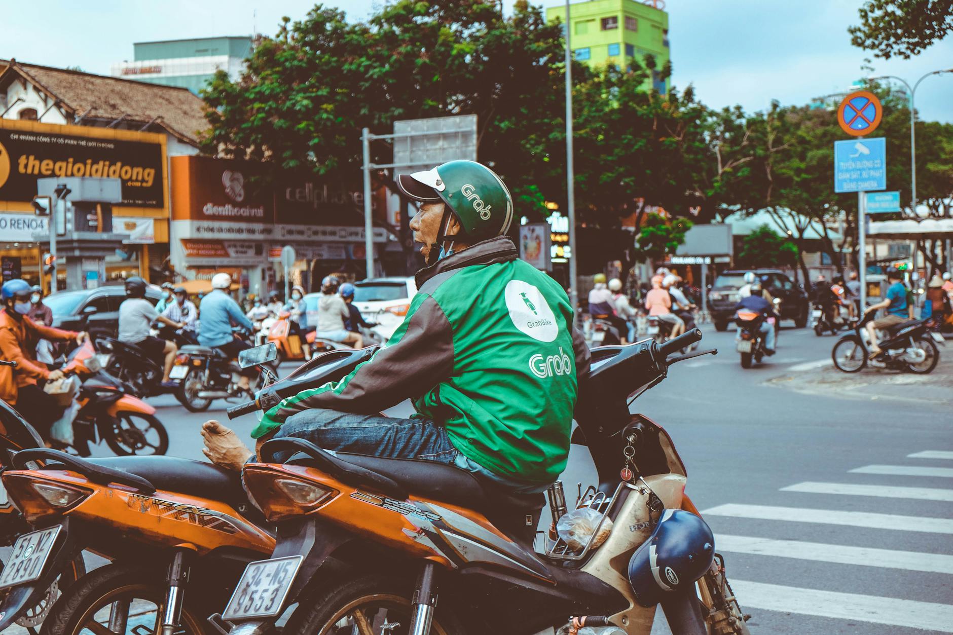 Motorbike rider in green jacket waits at a bustling urban intersection, surrounded by traffic.